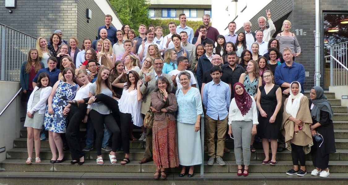 Etwa 60, vornehmlich junge Menschen, augenscheinlich unterschiedlicher Nationalitäten und Religionen, posieren herumalbernd für ein Gruppenfoto auf Treppenstufen.