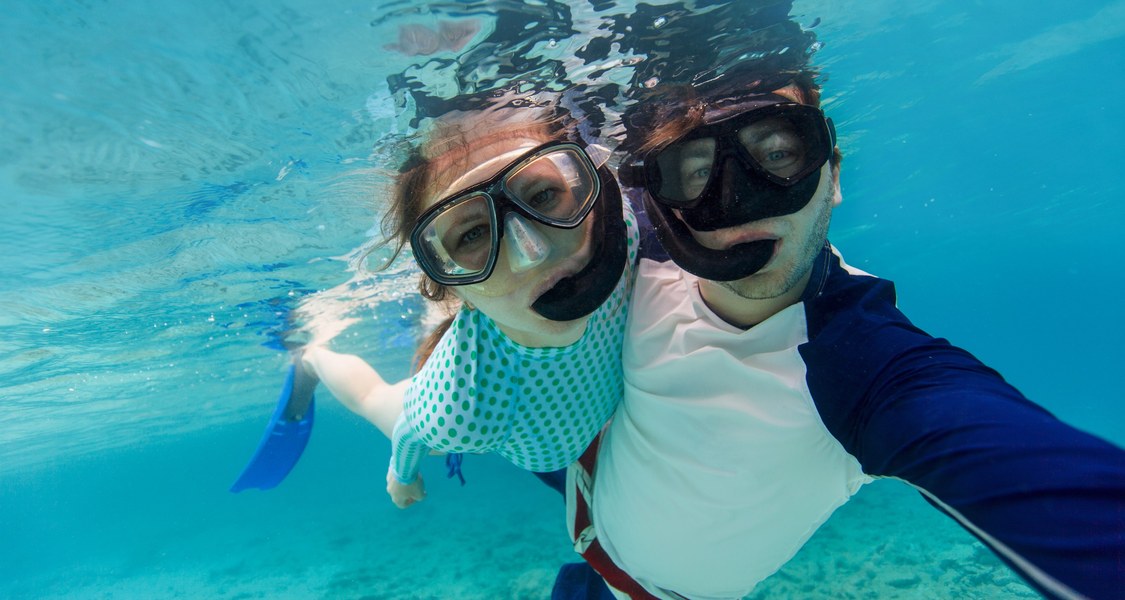 eine Frau und ein Mann mit normaler Straßenkleidung und mit Taucherbrille und Schnorchel schauen unter Wasser in die Kamera