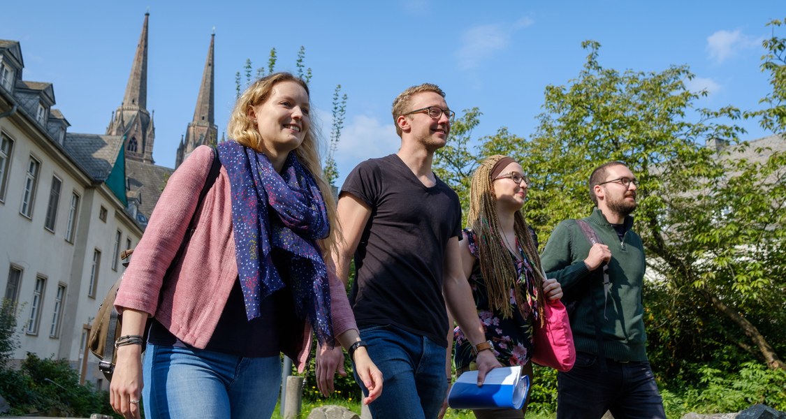 Drei Studierende sitzen unter einem Baum vor der Alten Universität und unterhalten sich. Sommerliche Szene