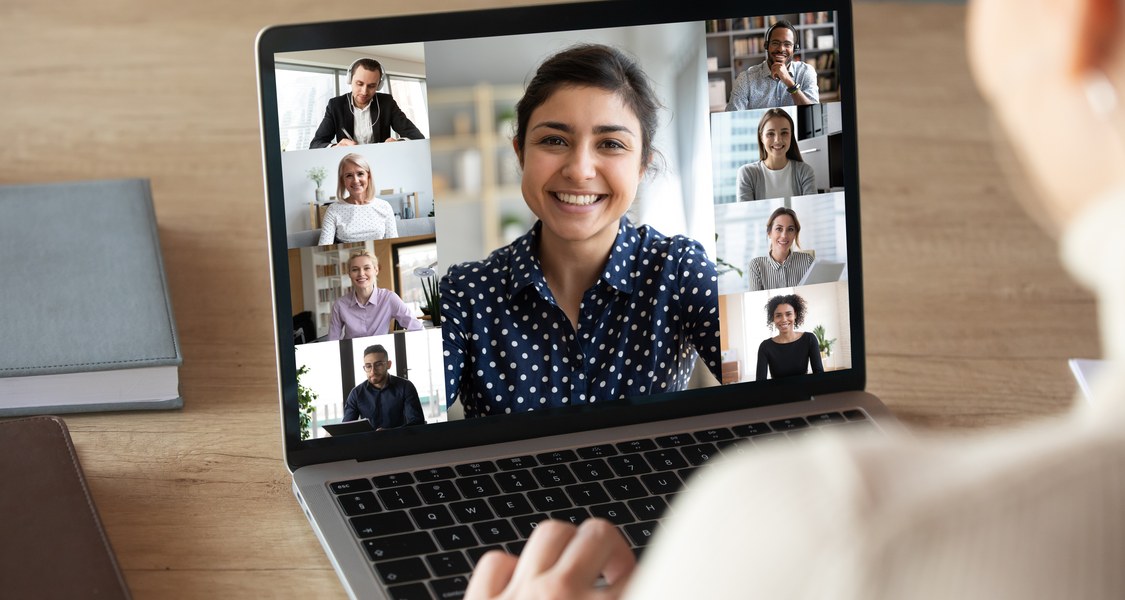 junge Frau sitzt vor Laptop, auf dessen Bildschirm verschiedene Gesichter in einer Collage wie bei einer Videokonferenz zu sehen sind