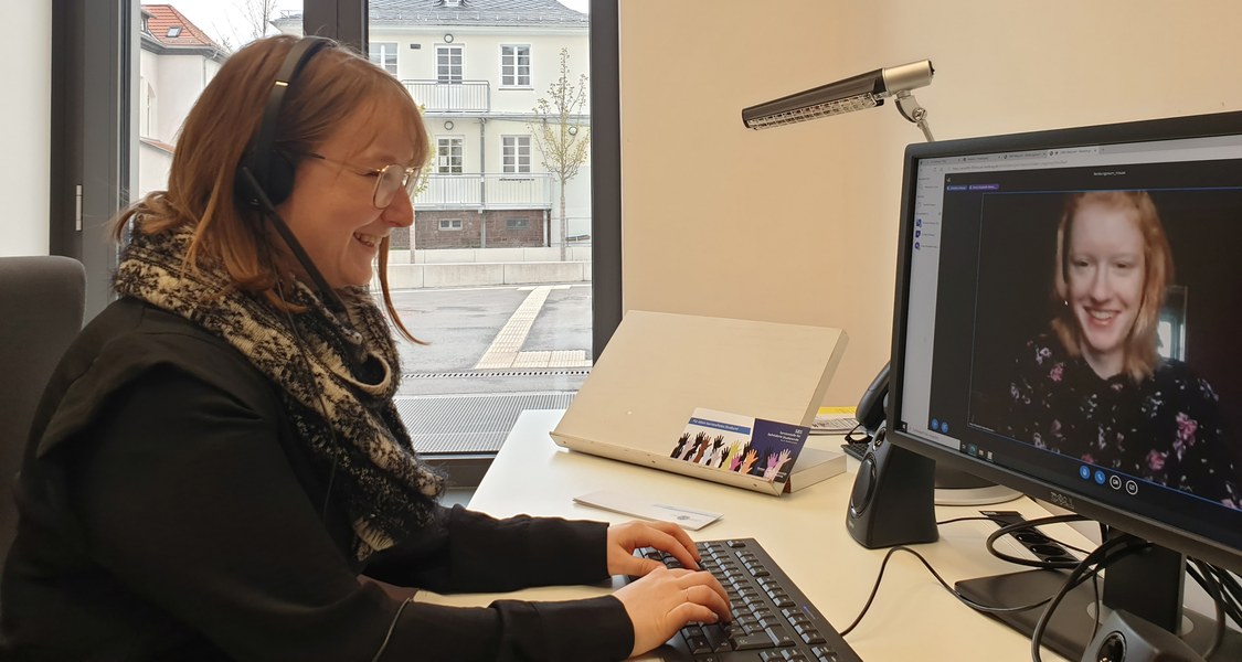 Frau mit Headset sitzt vor Bildschirm, auf dem eine andere Frau im Videocall zu sehen ist