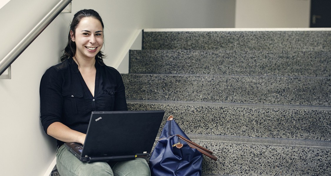 Lachende Studentin sitzt mit einem Laptop auf einer Treppe