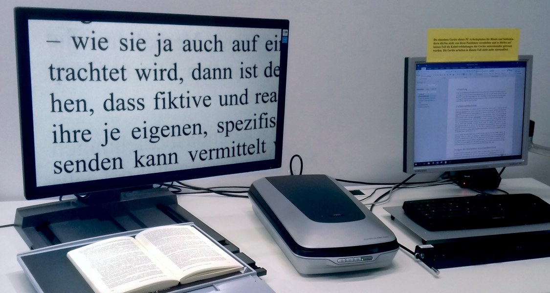 Auf dem Bild sind links ein Bildschirmlesegerät mit Kreuztisch und rechts ein Bildschirm mit Braillezeile und Tastatur zu sehen. Zwischen den beiden Geräten steht ein Flachbettscanner.