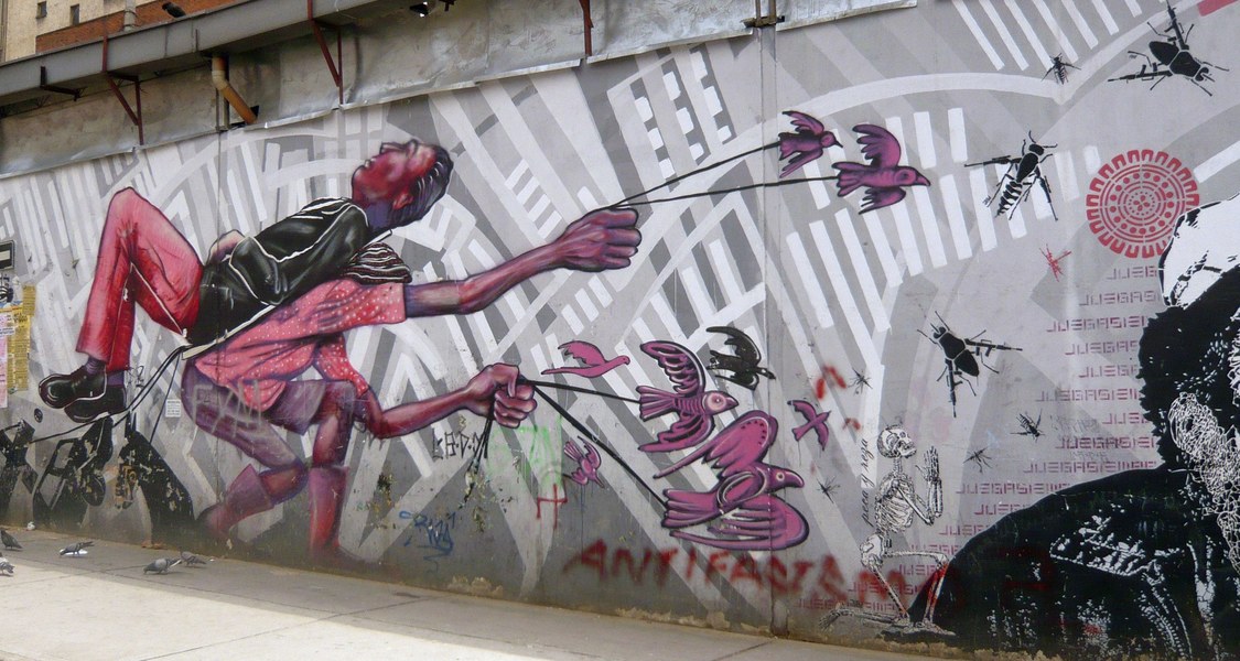 Das Wandbild zeigt zwei Personen, von denen eine mit angezogenen Beinen auf dem Rücken liegt. Die andere Person beugt sich unter dem Gewicht der ersten und hält mehrere Vögel an Leinen.