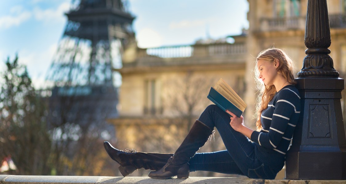 Junge Frau sitzt lesend auf einer Mauer, im Hintergrund Gebäude und der Pariser Eiffelturm.