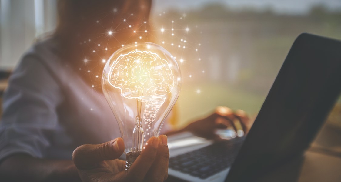 Leuchtende Glühbirne mit angedeuteter Gehirnstruktur, im Hintergrund sitzt eine Frau am Laptop.