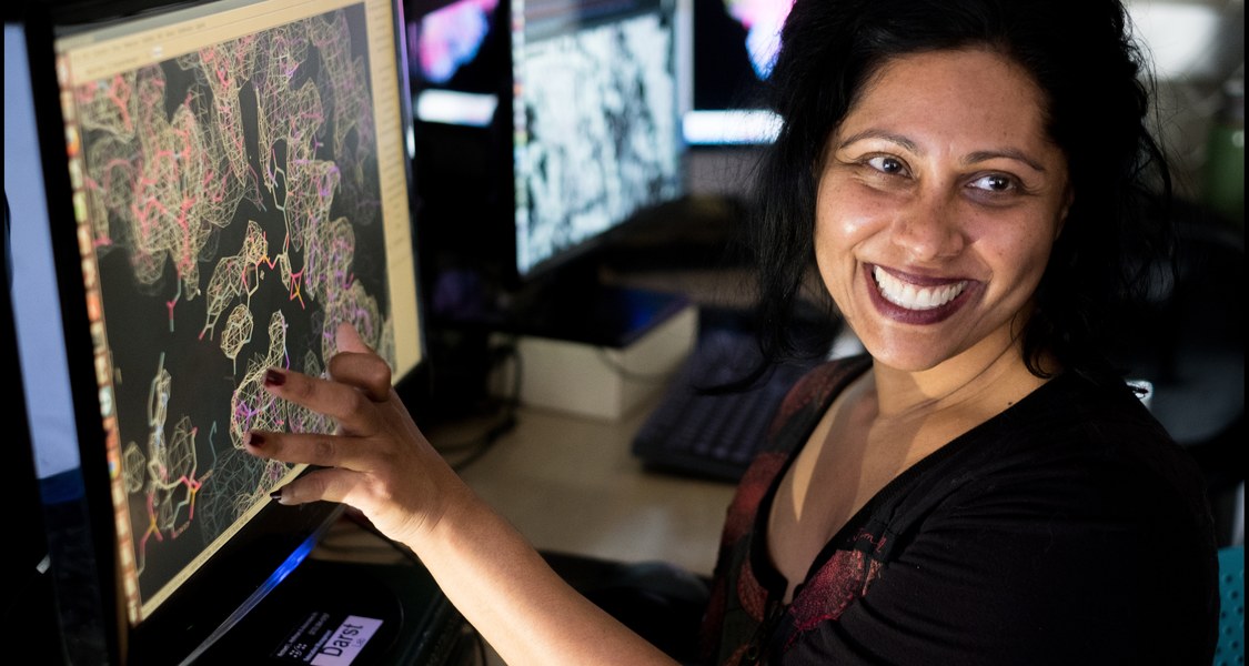 Forscherin zeigt mit der Hand auf einen Bildschirm und lächelt in die Kamera