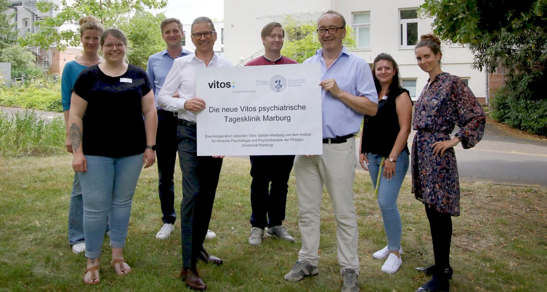 Gruppenfoto mit zwei Personen im Vordergrund, die ein Schild mit der Aufschrift "Die neue Vitos psychiatrische Tagesklinik Marburg" halten (links Michael Franz, rechts Winfried Rief)