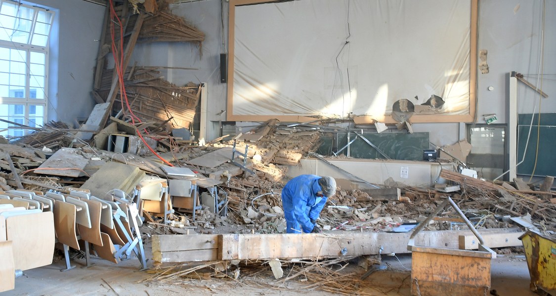 Ein Arbeiter ist mit Aufräumarbeiten im Hörsaal im Landgrafenhaus beschäftigt. Bestuhlung ist zusammengeschoben, ebenso herabgefallene Deckenteile, im Hintergrund Blick auf Leinwand und in den Dachstuhl des Gebäudes