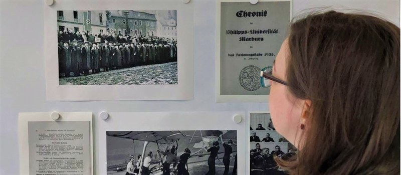 Halbprofilansicht einer Frau, die eine Wand mit Dokumenten und Schwarz-Weiß-Fotos betrachtet. Auf den Fotos sind Szenen aus der Zeit von 1933-1945 zu sehen.