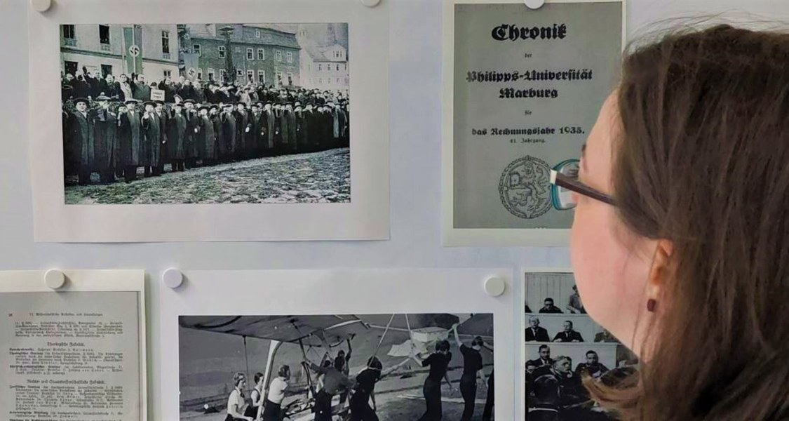 Halbprofilansicht einer Frau, die eine Wand mit Dokumenten und Schwarz-Weiß-Fotos betrachtet. Auf den Fotos sind Szenen aus der Zeit von 1933-1945 zu sehen.
