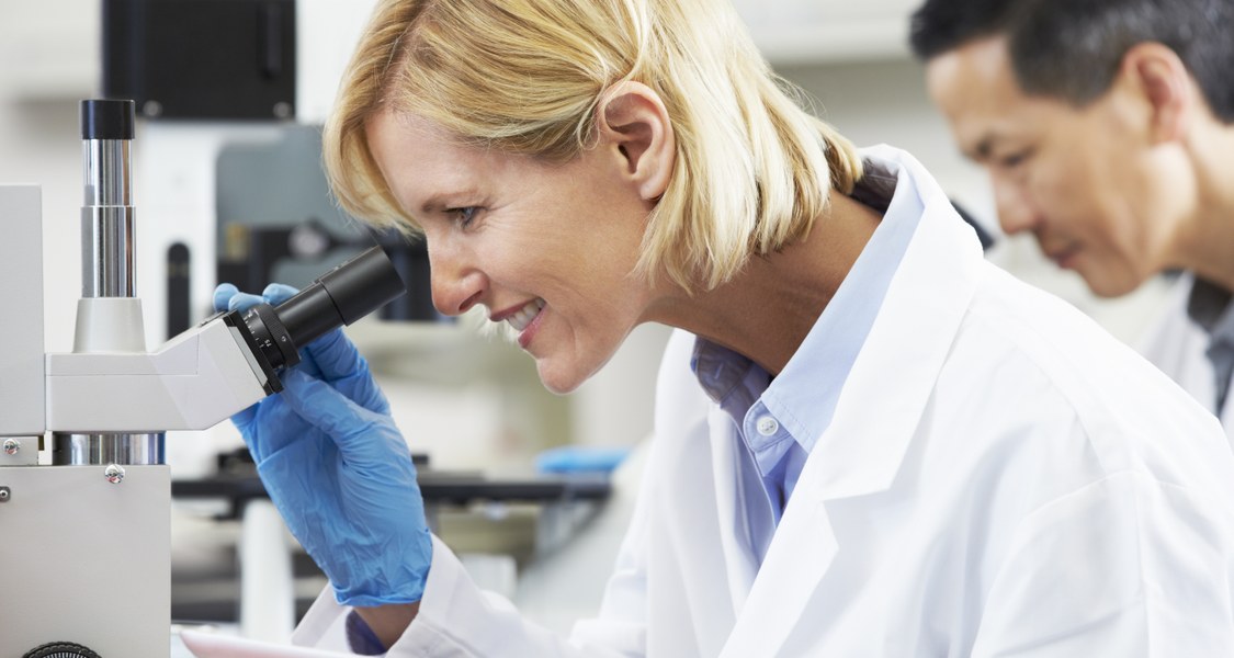 Blonde Frau in Laborkittel blickt in ein Mikroskop