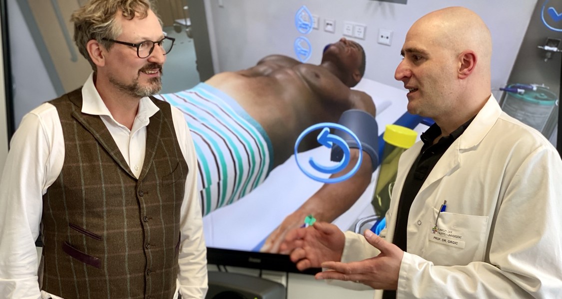 zwei Personen vor einem großen Bildschirm, der einen virtuellen Patienten zeigt