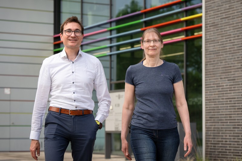 beide Personen laufen auf die Betrachter zu vor dem Hintergrund des Haupteingangs des Gebäudes von Synmikro.