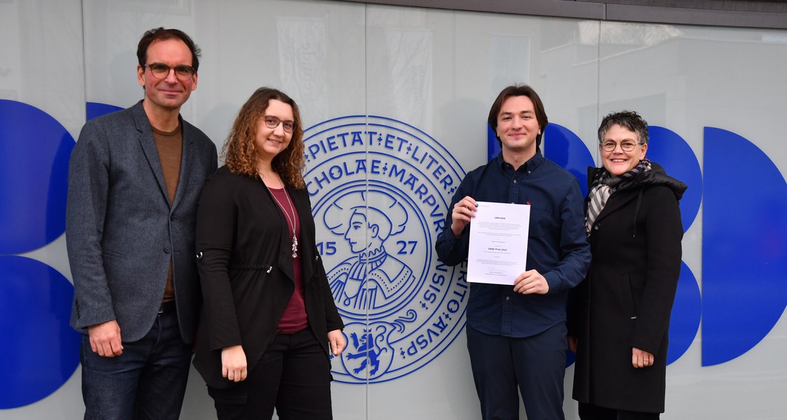 Vier Menschen vor dem Uni-Hauptgebäude, ein Mensch hälte eine Urkunde in die Kamera