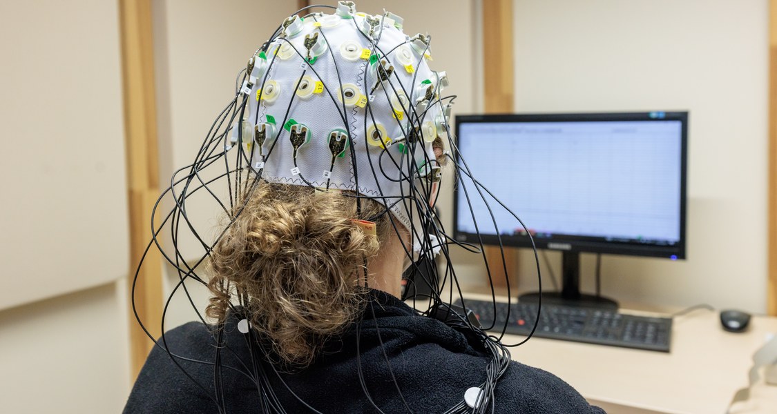 Frau mit EEG-Haube von hinten, schaut auf einen Bildschirm, auf dem ein Elektroenzephalogramm zu sehen ist