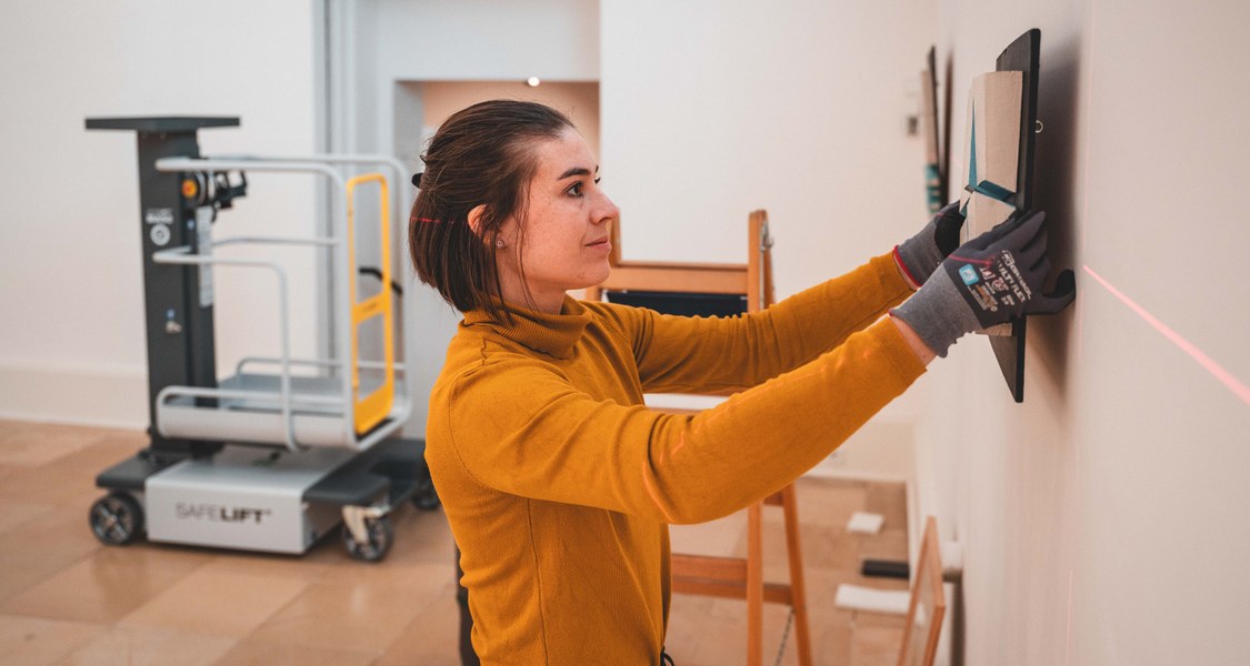 Eine Frau in gelebem Pullover hängt ein Kunstwerk an die Wand.