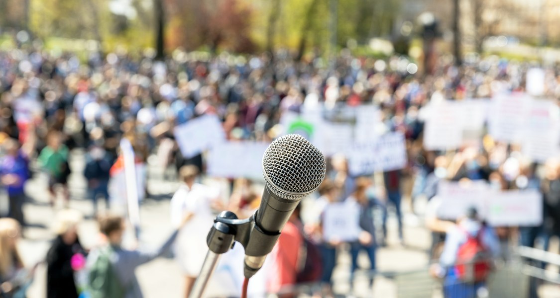 Symboldbild Demokratieforschung: Ein Mikrofon in Nahaufnahme, im Hintergrund eine unscharfe Menschenmenge