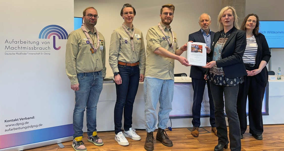 Roll-up mit Aufschrift "Aufarbeitung von Machtmissbrauch. Deutsche Pfadfinder*innenschaft St. Georg", daneben Personen mit Pfadfinder*innen-Hemd gekleidet. Eine Frau übergibt einer dieses Personen ein Dokument. Zwei weitere Personen im Hintergrund