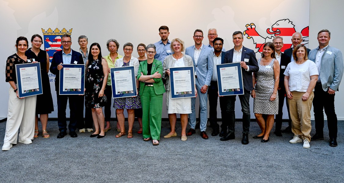 Gruppenfoto mit 20 Personen, 5 Personen zeigen große Urkunden u.a. mit dem Schriftzug LOEWE, im Hintergrund sind teilweise Logos mit dem hessischen Löwen zu sehen