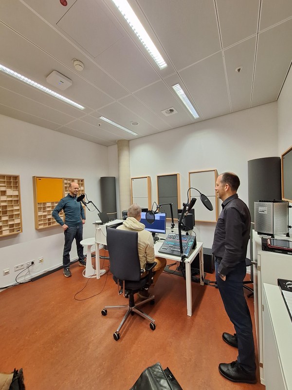 im Tonstudio stehen zwei Menschen links und rechts vor Mikrofon, in der Mitte ein Mensch am Mischpult