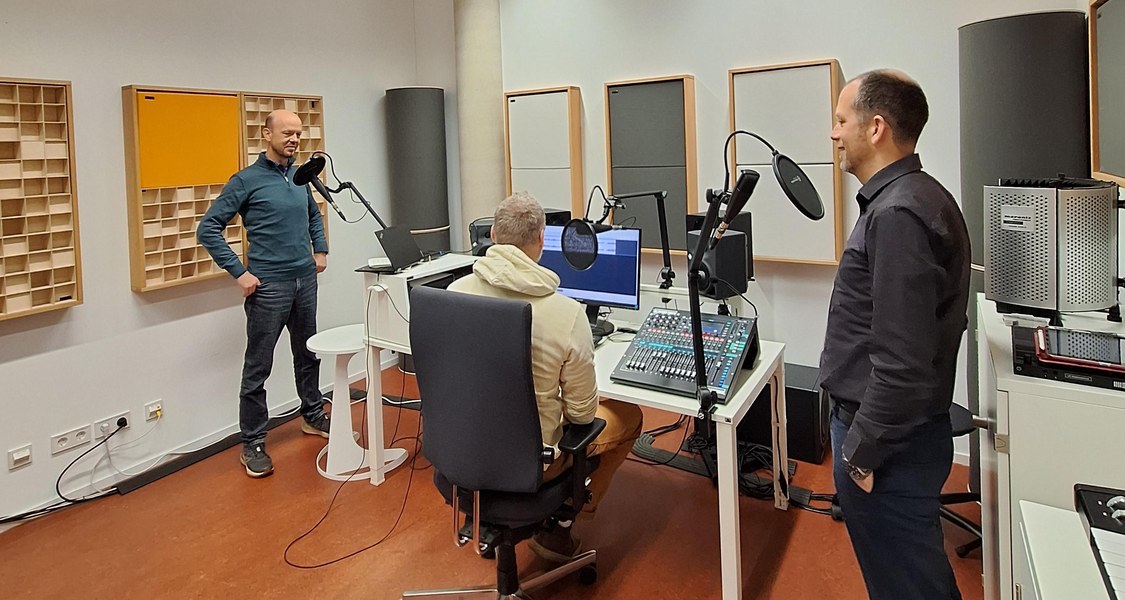 im Tonstudio stehen zwei Menschen links und rechts vor Mikrofon, in der Mitte ein Mensch am Mischpult