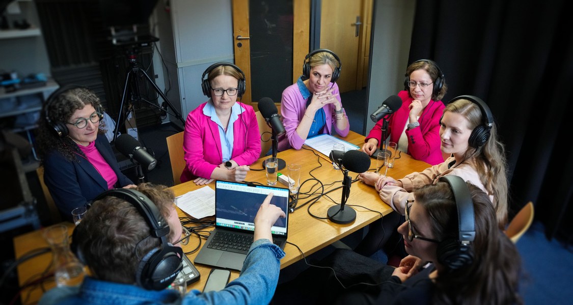 Gruppenfoto von fünf Personen mit Kopfhörern und Mikrofonen in einem Podcast-Studio