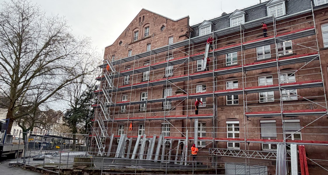 Fassade mit Gerüst davor, im Vordergrund Baumaterial, ein Bauzaun und weitere Gegenstände auf dem Parkplatz