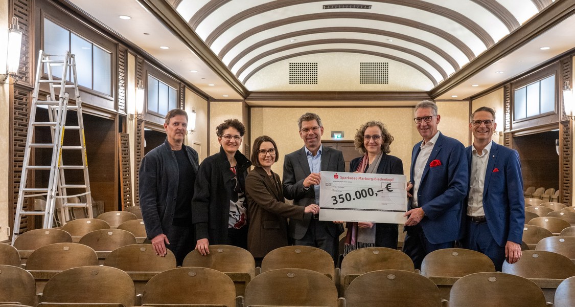 Sieben Personen stehen zwischen Besucherstühlen im Musiksaal und halten einen Scheck