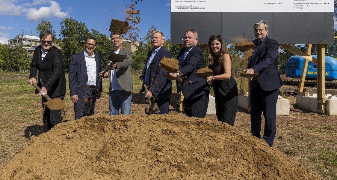 Die abgebildeten Personen haben Spaten in der Hand und werfen symbolisch Sand nach oben. Im Hintergrund steht das Bauschild für den Bau der neuen Menseria.