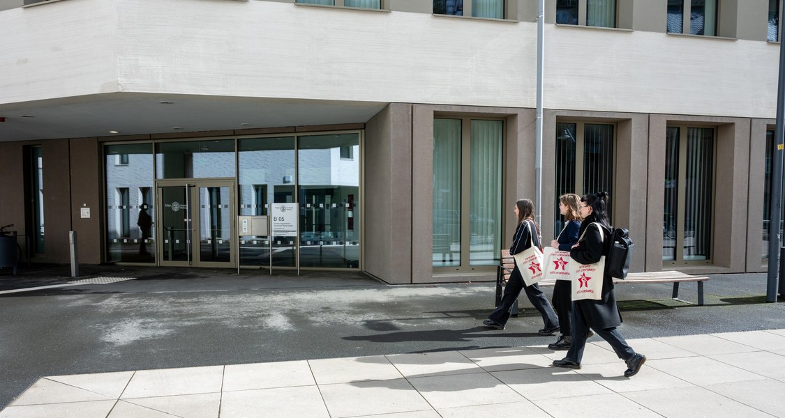 Eingang des Gebäudes Sprachatlas mit Fassade, drei Personen mit Stofftaschen laufen von rechts auf Eingang zu
