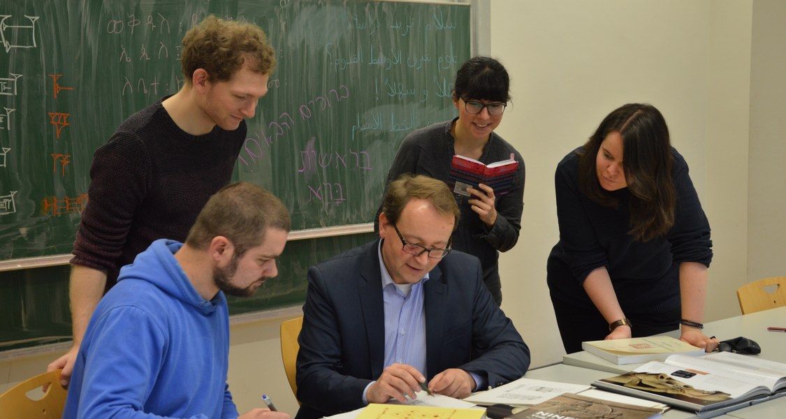 Der Professor sitzt vor einem Tische voller Fachbücher, von Studierenden umgeben