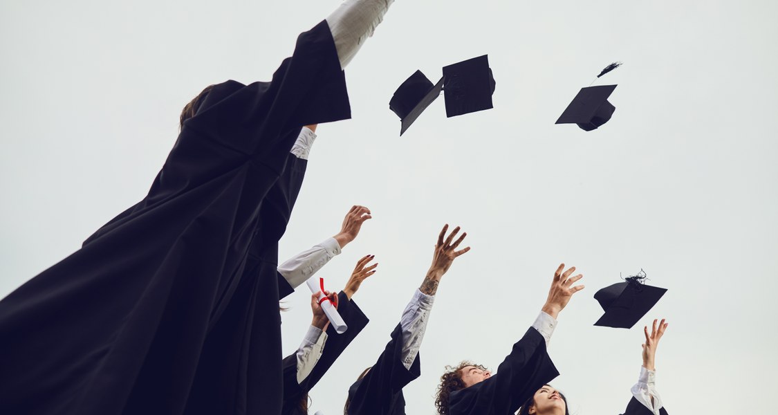 Auf dem Bild sind mehrere Personen in einer Abschlussrobe zusehen, die ihre Hüte in die Luft werfen.