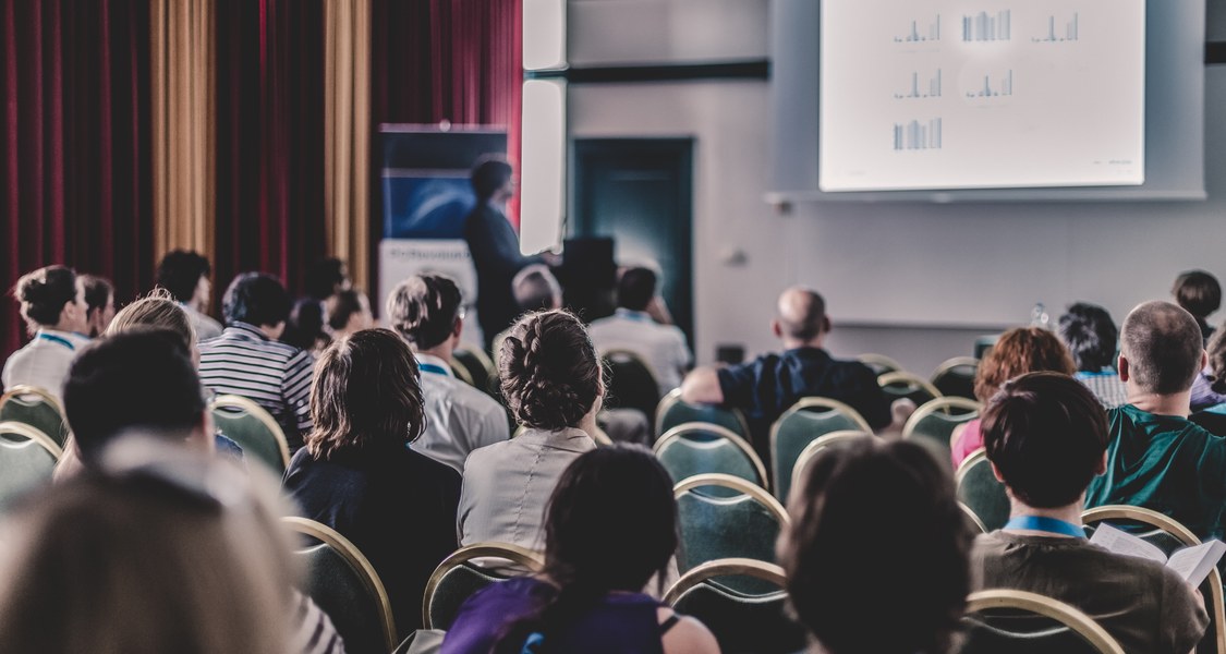 Vortragsraum von hinten. Viele Menschen sitzen im Publikum. Von diesen ist nur der Hinterkopf zu erkennen. Vorne wird ein Vortrag mit einer Powerpoint Präsentation gehalten.