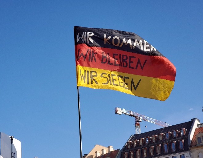 Man sieht eine Deutschlandflagge an einem Fahnenmast. Auf die schwarz-weiß-roten Streifen der Flagge wurde gut sichtbar in Großbuchstaben "Wir kommen Wir bleiben Wir siegen" geschrieben.