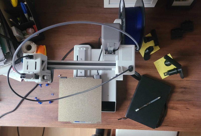 Draufsicht auf einen Schreibtisch. Im Fokus steht ein 3D-Drucker für den Privatgebrauch, daneben liegt ein geschlossenes Notizbuch mit einem Schreibtisch.