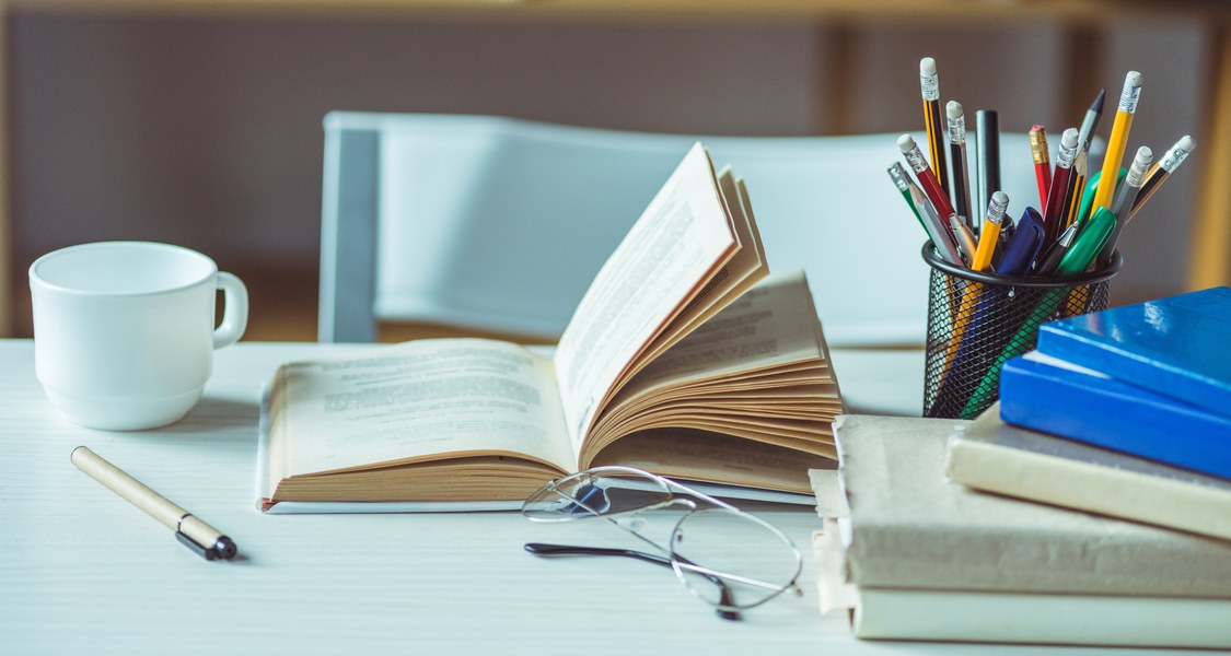 Auf dem Foto ist ein Arbeitsplatz mit Büchern, Stiften und einer Tasse abgebildet.