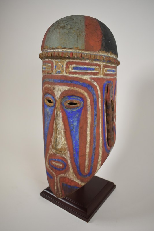 Bemalte Maske mit Gucklöcher für die Augen; Mund, Nase und Ohren werden angedeutet. Holz aus einem Stück geschnitzt, bemalt in rot, weiß, blau und grau-schwarz.