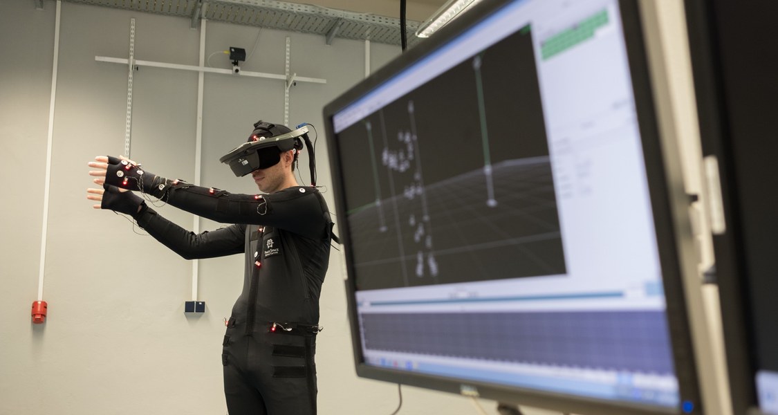 Eine Person in einem Motion Capture Anzug und Virtual Reality Brille greift nach virtuellen Boxen. Im Vordergrund sieht man einen Monitor mit Motion Capture Software.