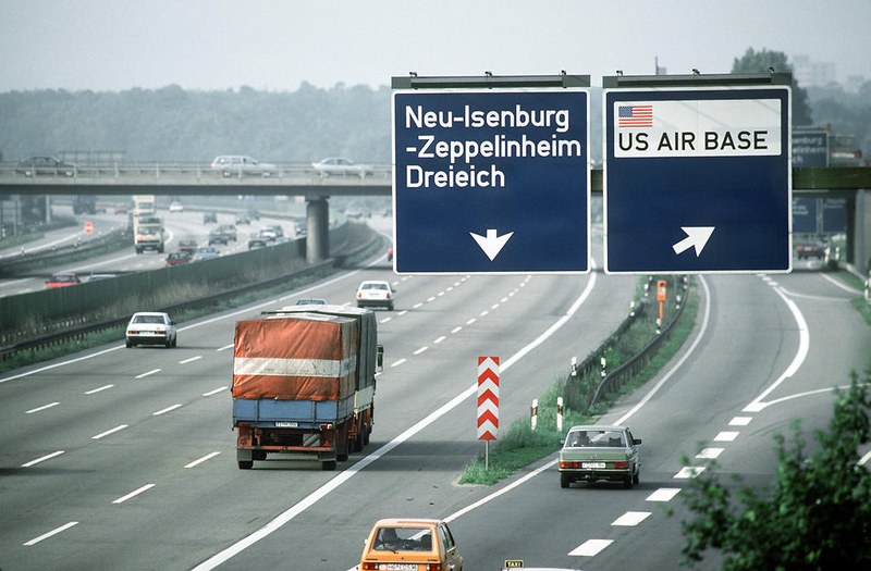 Mehrspurige Autobahn mit Verkehr. Im Vordergrund hängen zwei große blaue Verkehrsschilder: Links führt ein Pfeil nach unten für „Neu-Isenburg -Zeppelinheim Dreieich“, rechts weist ein Pfeil nach rechts mit US-Flagge auf die „US AIR BASE“ hin.