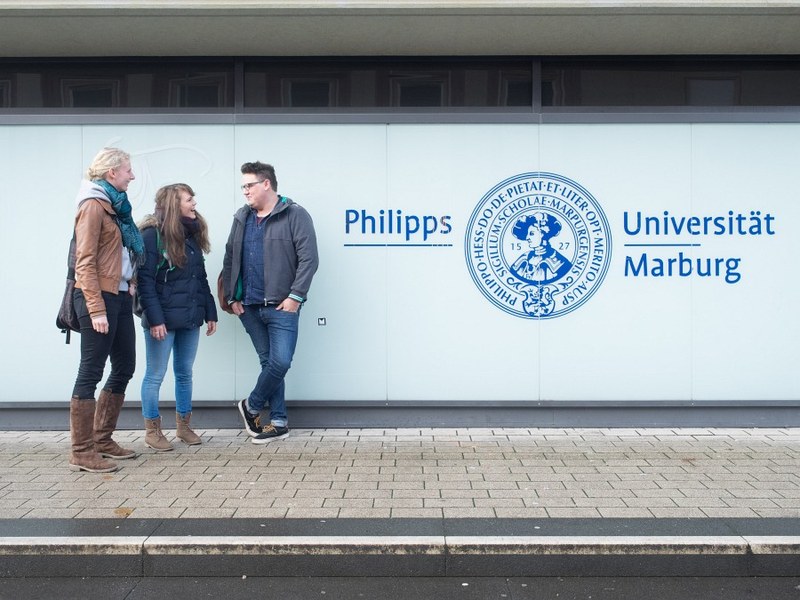 Drei Studierende neben dem Logo der Uni Marburg am Hauptgebäude in der Biegenstraße.jpg