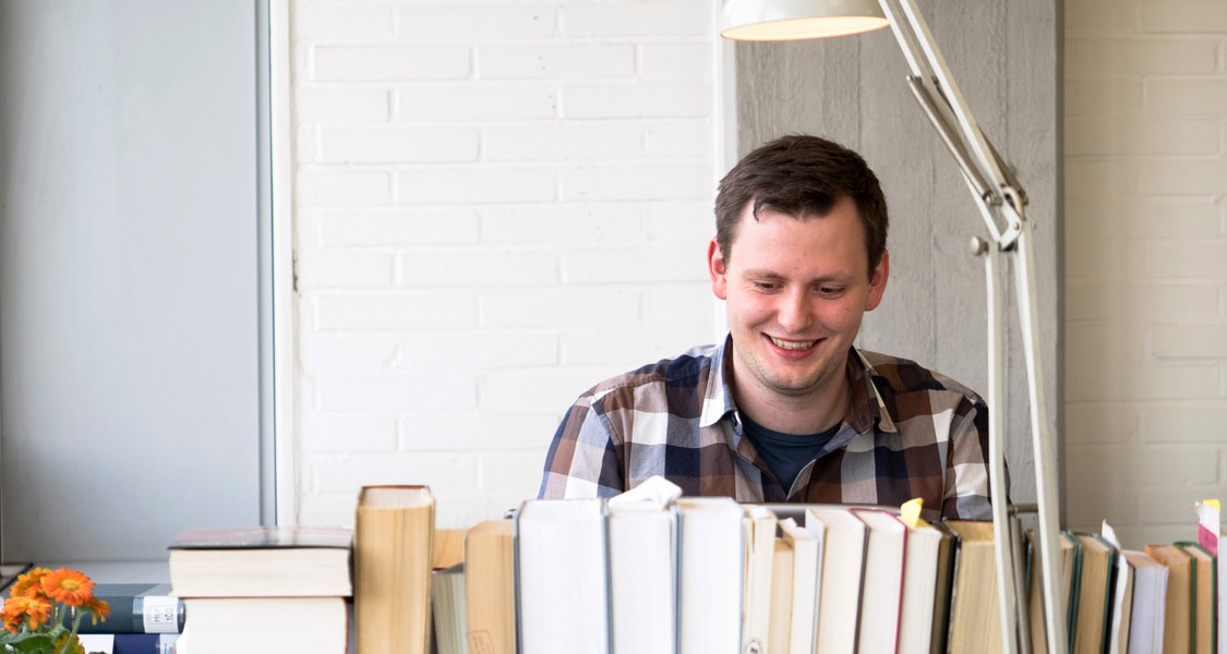 Studierender unter einer Lampe an einem Arbeitsplatz mit einer langen Reihe Bücher ringsherum.