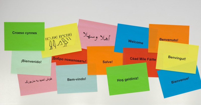 Mehrere bunte Zettel auf Whiteboard mit Aufdruck "Willkommen" in verschiedenen Sprachen.