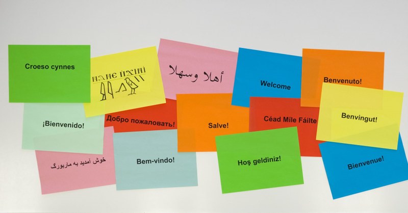 Mehrere bunte Zettel auf Whiteboard mit Aufdruck "Willkommen" in verschiedenen Sprachen.