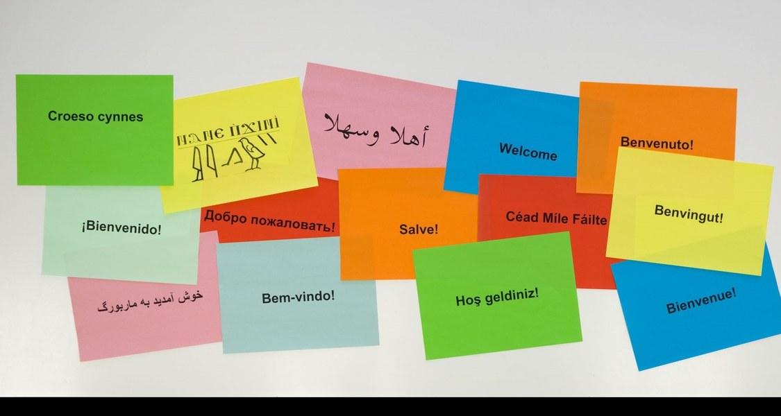 Mehrere bunte Zettel auf Whiteboard mit Aufdruck "Willkommen" in verschiedenen Sprachen.