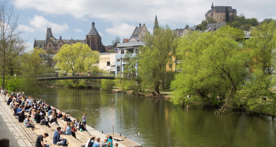 Studierende sitzen auf den Lahnterrassen, der Blick geht über die Lahn und die Mensabrücke und Marburgs Oberstadt mit der Alten Universität links und dem Schloss oben rechts im Bild