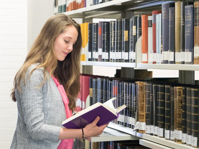 Studierende liest ein Buch vor einem Bibliotheksregal.