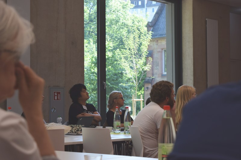 Gruppe von Tagungsteilnehmern an den Tischen verfolgt einen Vortrag. Im Hintergrund der Blick aus dem Fenster auf Bäume des alten Botanischen Gartens, ein altes Universitätsgebäude sowie weiter oben auf Häuser der Marburger Oberstadt.