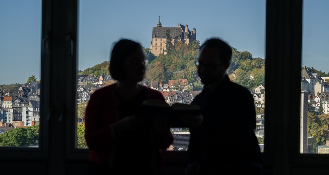 Zwei nicht erkennbare Personen vor einem Bürofenster mit Blick auf das Marburger Schloss im Fokus