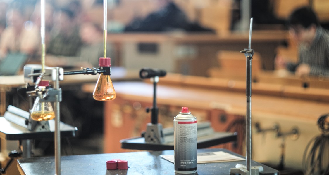 ein Versuchsaufbau mit Stativ, Kolben, Klemmen, im Hintergrund unscharf Hörsaalstuhlreihen mit Publikum